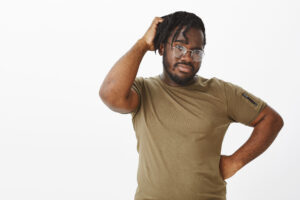 Indoor shot of unsure handsome african american in glasses, holding hand on hip and scratching head, being doubtful while thinking, making choice with confused expression, standing in hesitations